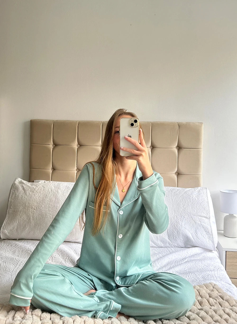 Woman taking a mirror selfie wearing light blue pajamas in a bedroom.