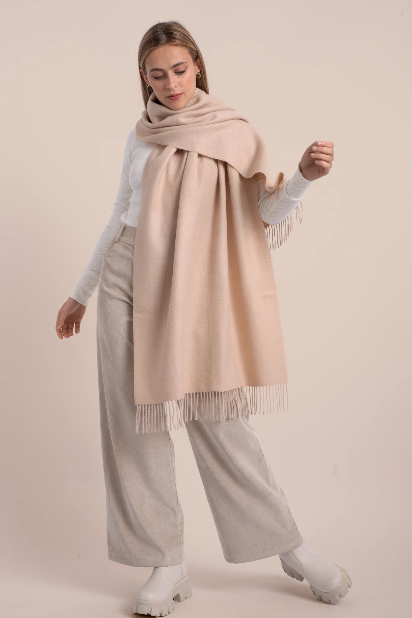 Woman wearing a beige scarf with fringe, light gray sweater, and wide-leg pants on a beige background