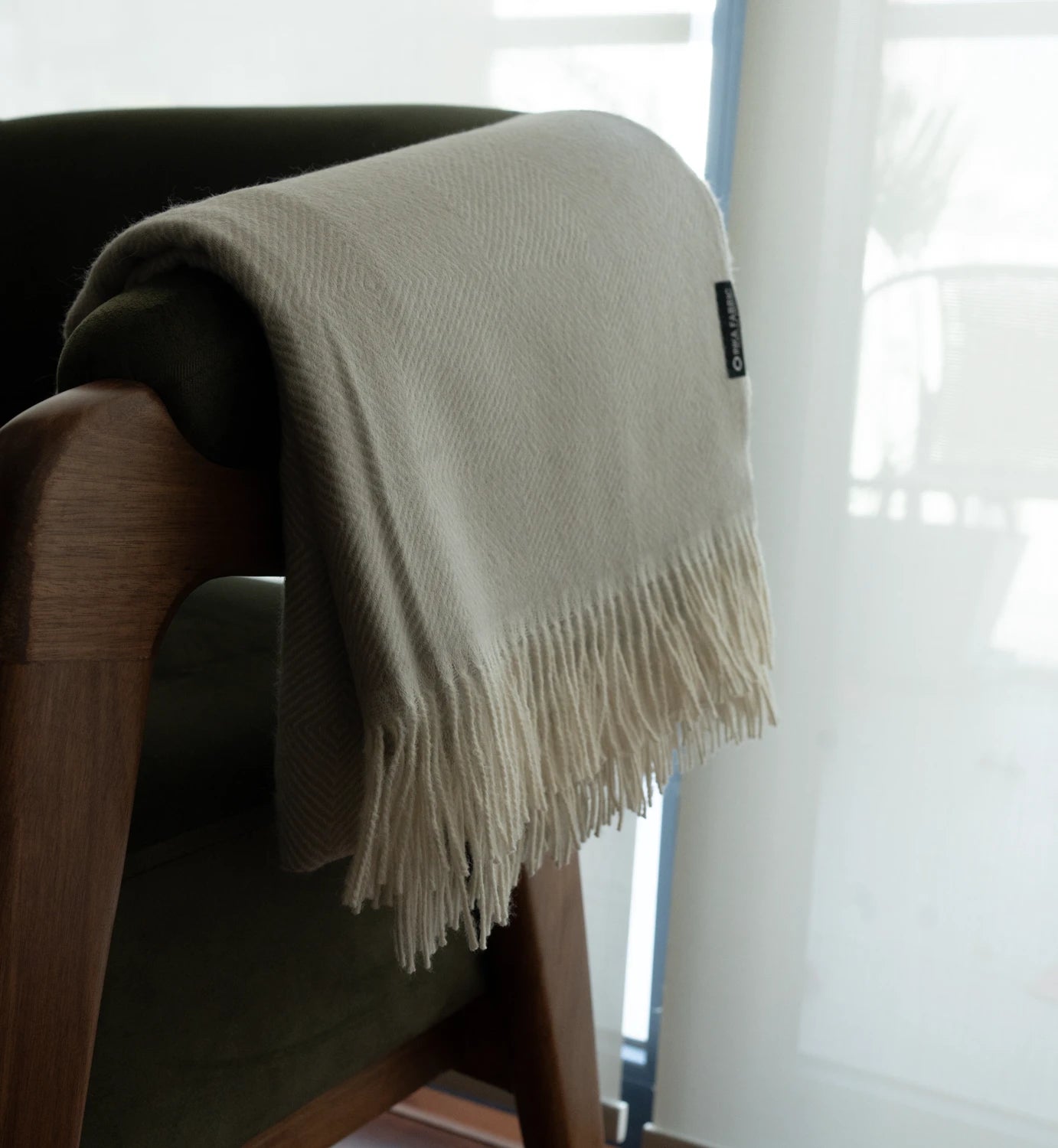 Beige throw blanket with fringe draped over a wooden chair in a room with a window.