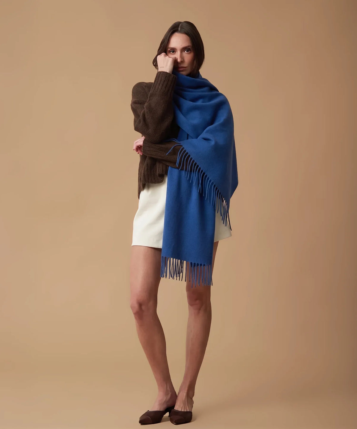 A model wearing a vibrant blue fringed wrap shawl over a brown knit sweater, white shorts, and brown flat shoes, posed against a solid beige studio background.