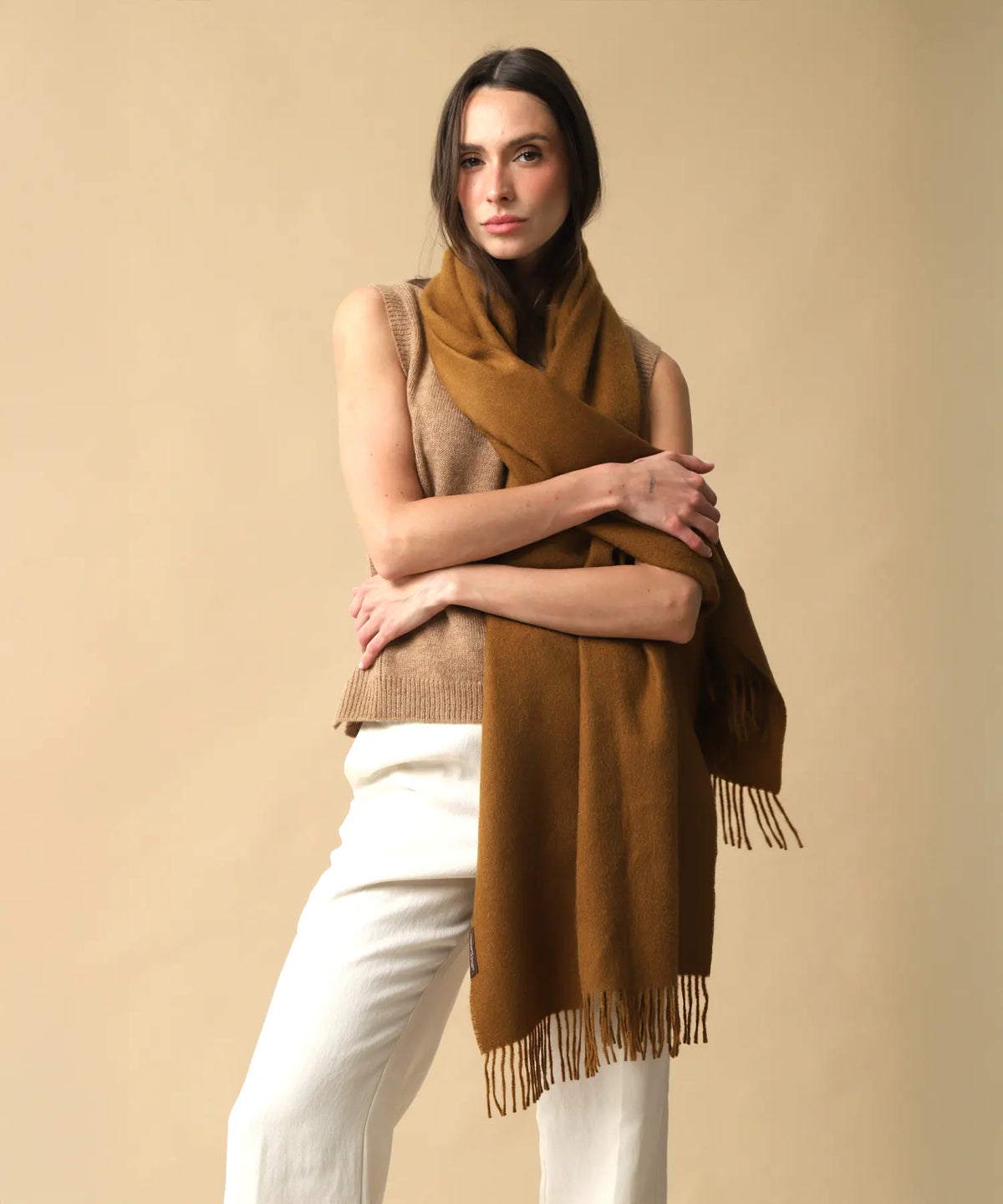 A woman modeling a warm brown woolen scarf wrapped around her shoulders, paired with a beige knit vest and white pants, set against a solid beige studio background.