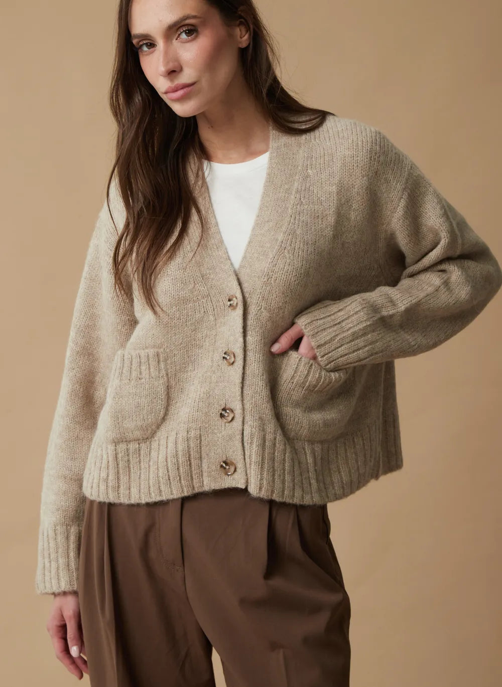 A woman wearing a beige chunky knit cardigan with brown buttons, layered over a white top and paired with brown trousers, set against a solid beige background.