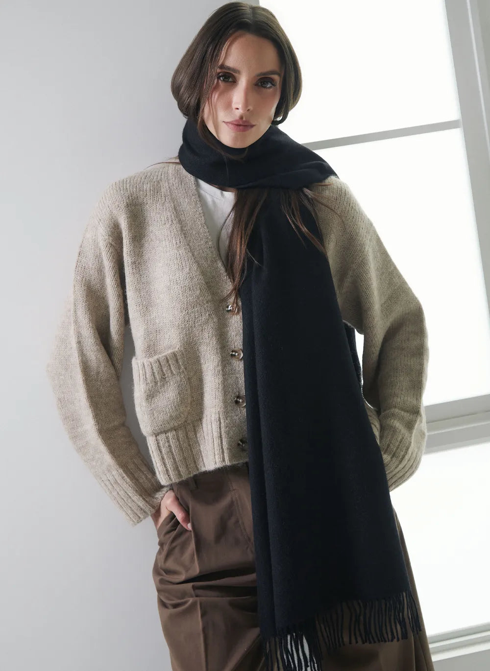 A woman wearing a beige knit cardigan, a dark navy scarf, and brown trousers, standing in front of a bright window.