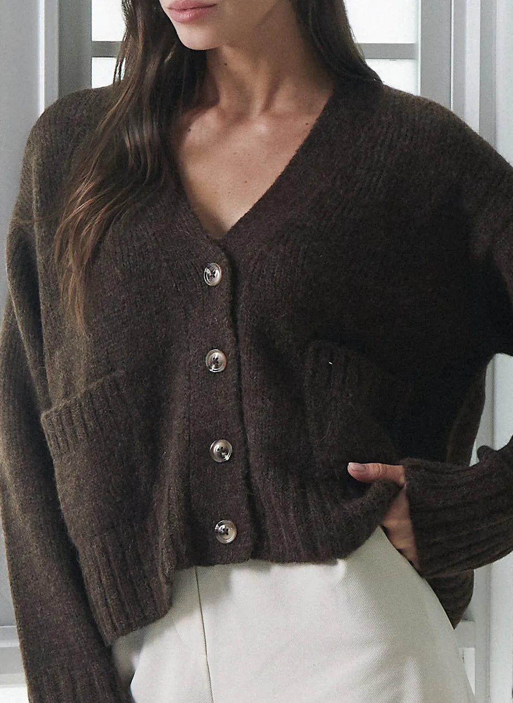 A woman wearing a dark brown knit cardigan sweater with silver buttons and a V-neck, paired with white pants.