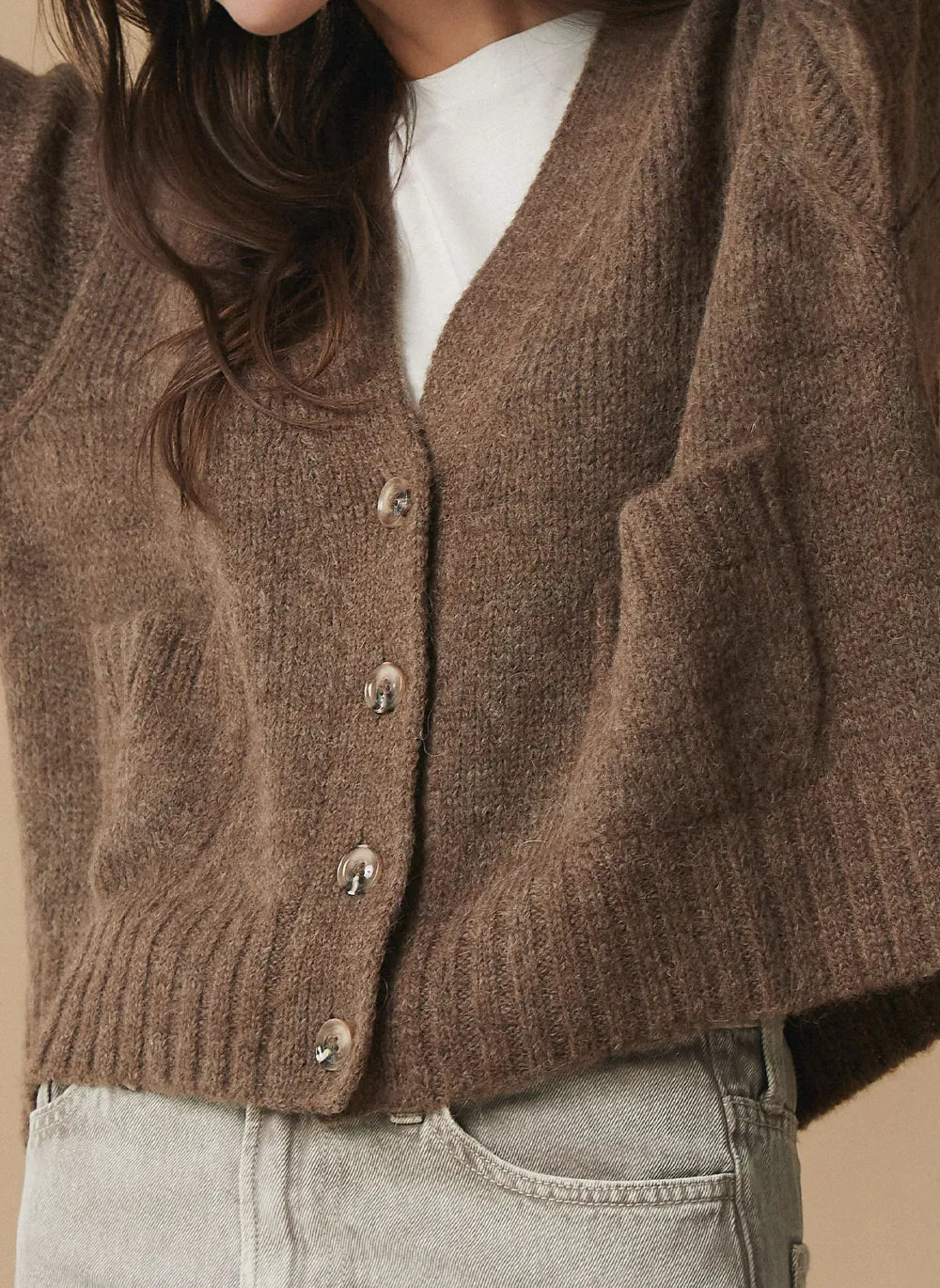 A close-up of a person wearing a brown chunky knit cardigan with front buttons over a white top and light gray jeans.