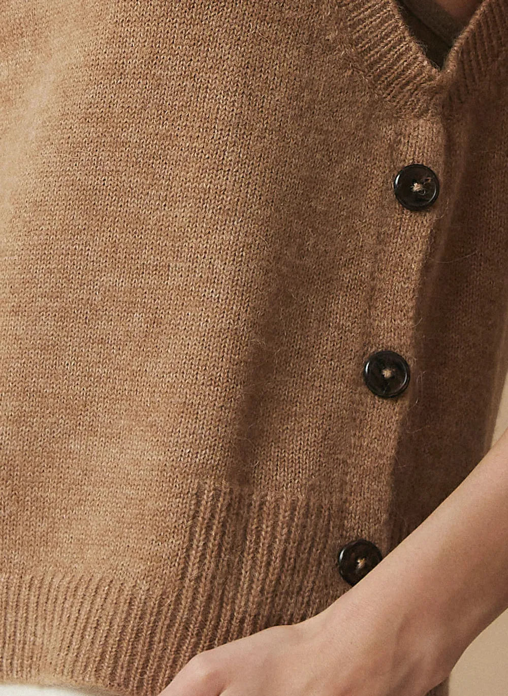 Close-up of a brown knit cardigan sweater with dark buttons, worn by a model whose hand is visible at the bottom right.