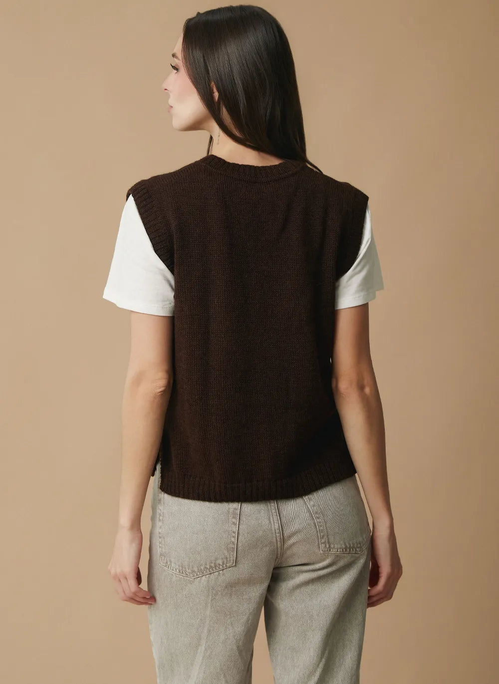 A woman shown from the back wearing a dark brown sleeveless knit vest over a white short-sleeve t-shirt and light gray pants, set against a solid beige studio background.