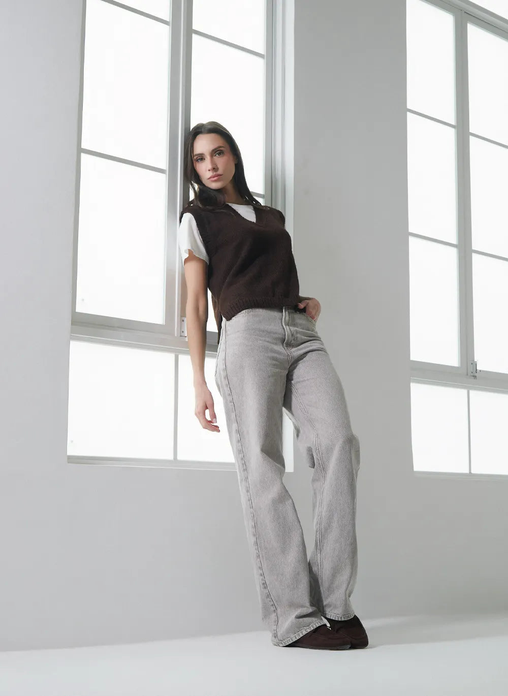 A woman modeling light gray wide-leg jeans, a dark brown sleeveless vest over a white t-shirt, and dark brown shoes, standing in a bright white room with large windows.