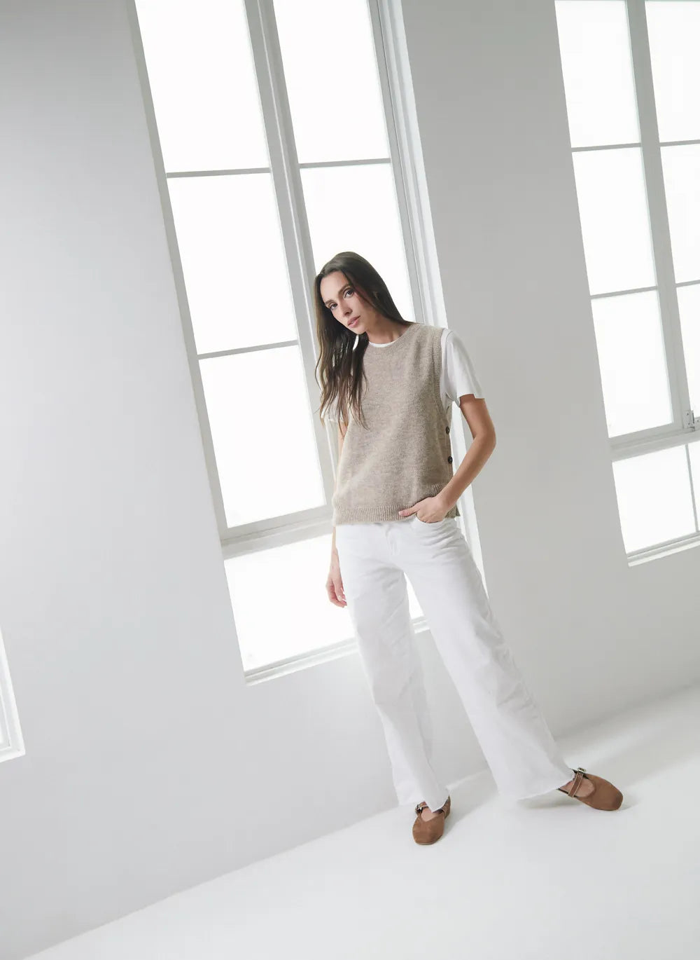 A woman stands in a bright, minimalist white room with tall windows, wearing a beige knit sleeveless vest over a white t-shirt, white wide-leg pants, and brown flat shoes.