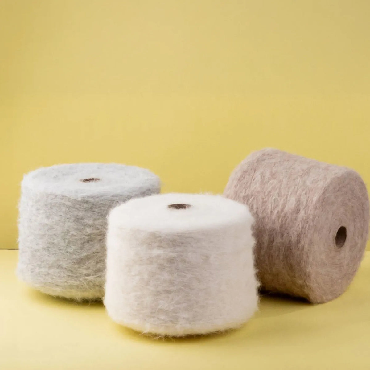 Three fuzzy yarn or fabric spools in light gray, off-white, and taupe on a solid pale yellow background.