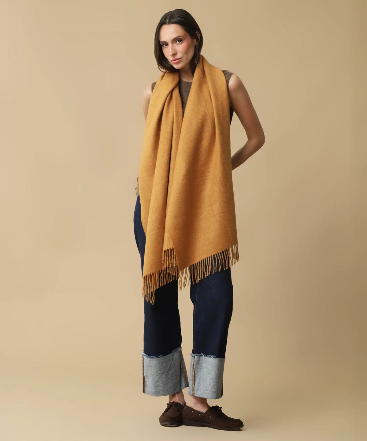 A woman modeling a thick mustard yellow fringed scarf wrapped around her shoulders, paired with dark blue jeans, brown loafers, and a dark gray top, against a solid beige studio background.