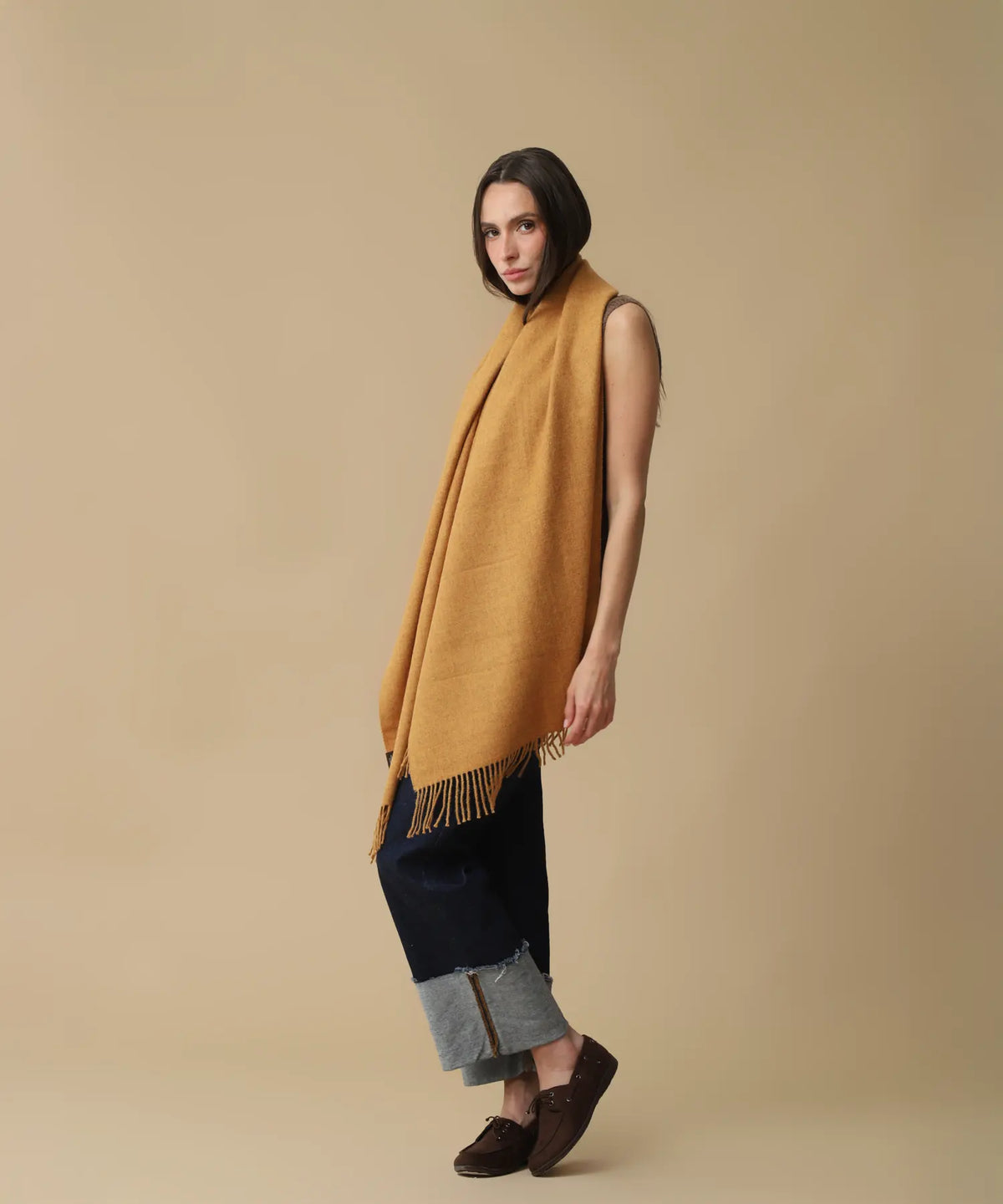 A woman modeling a mustard yellow knit shawl with fringe trim, styled over a dark navy sleeveless top and cuffed jeans, against a solid beige studio background.