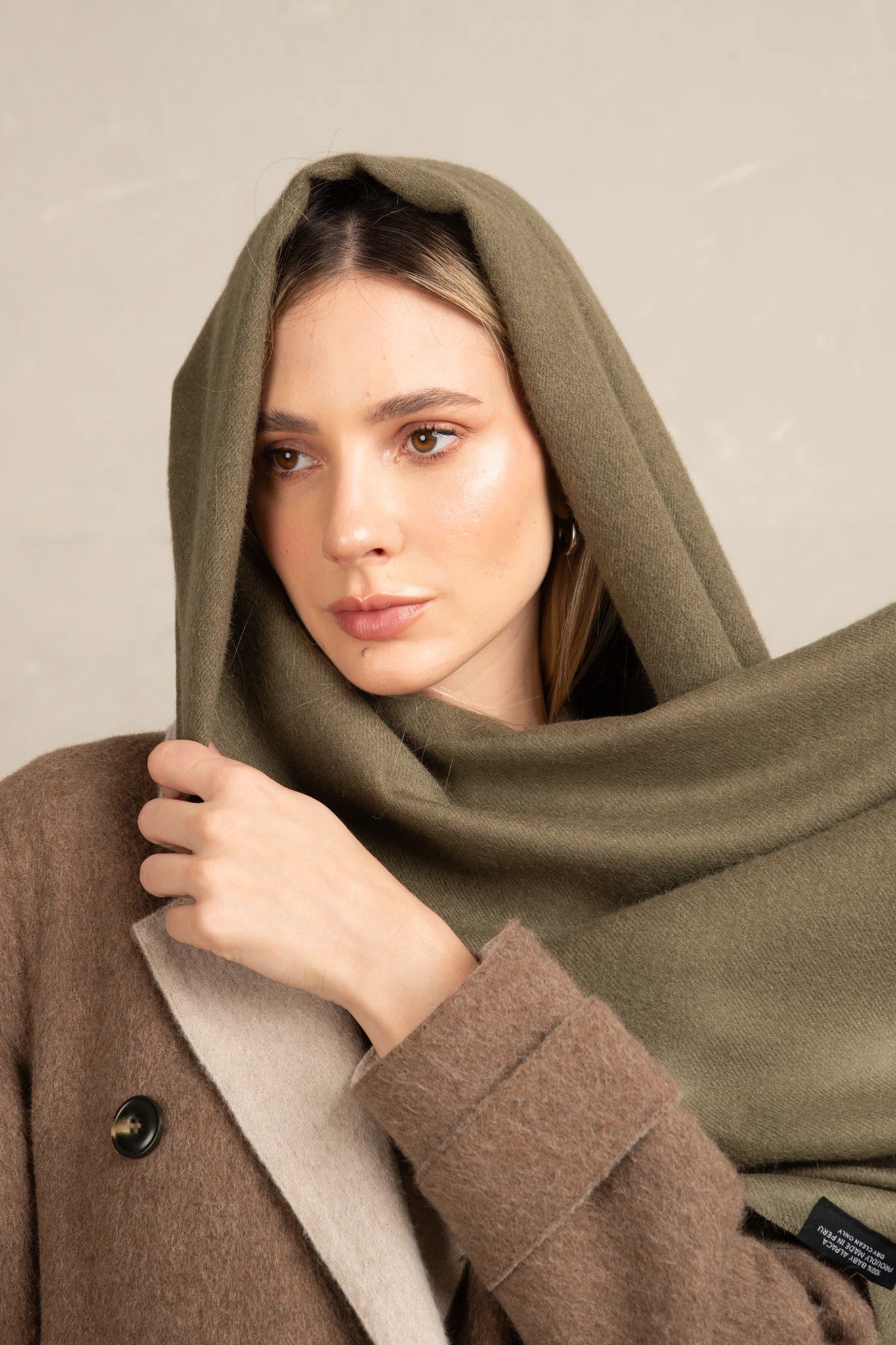 Woman wearing a brown coat with a green scarf draped over her head against a neutral background