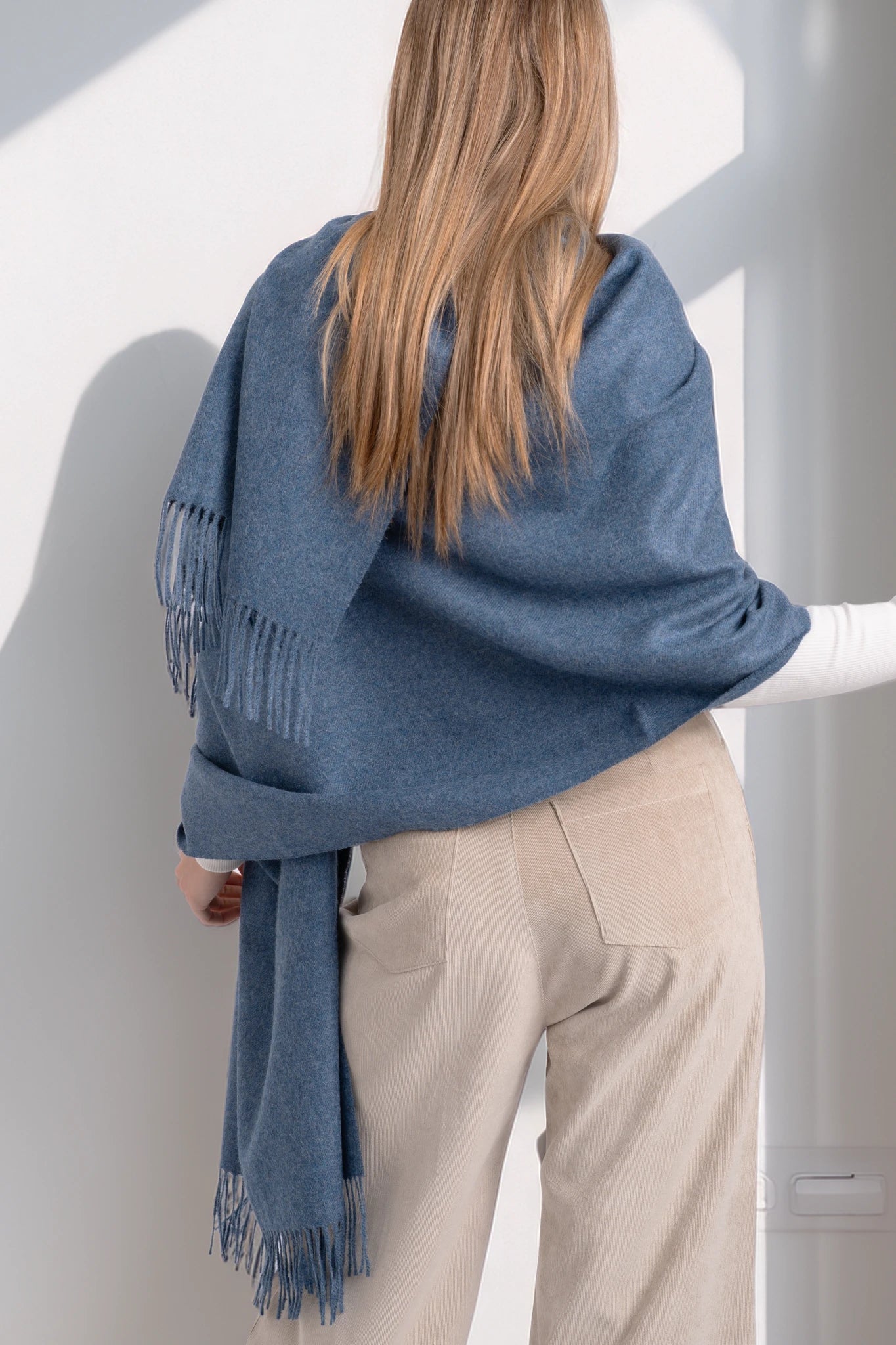 Person wearing a blue scarf with fringe, standing against a white wall.