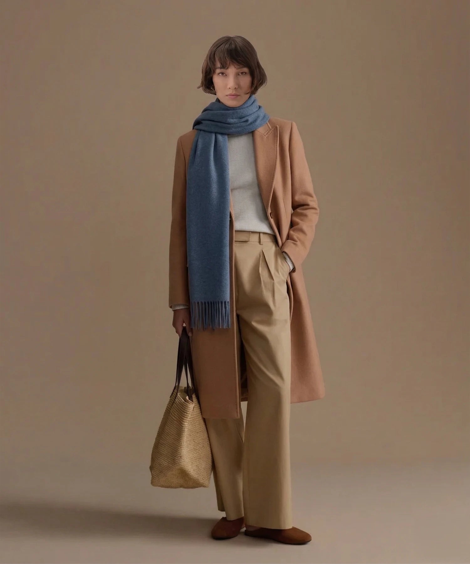 Woman wearing a brown coat, beige pants, and a blue scarf against a beige background