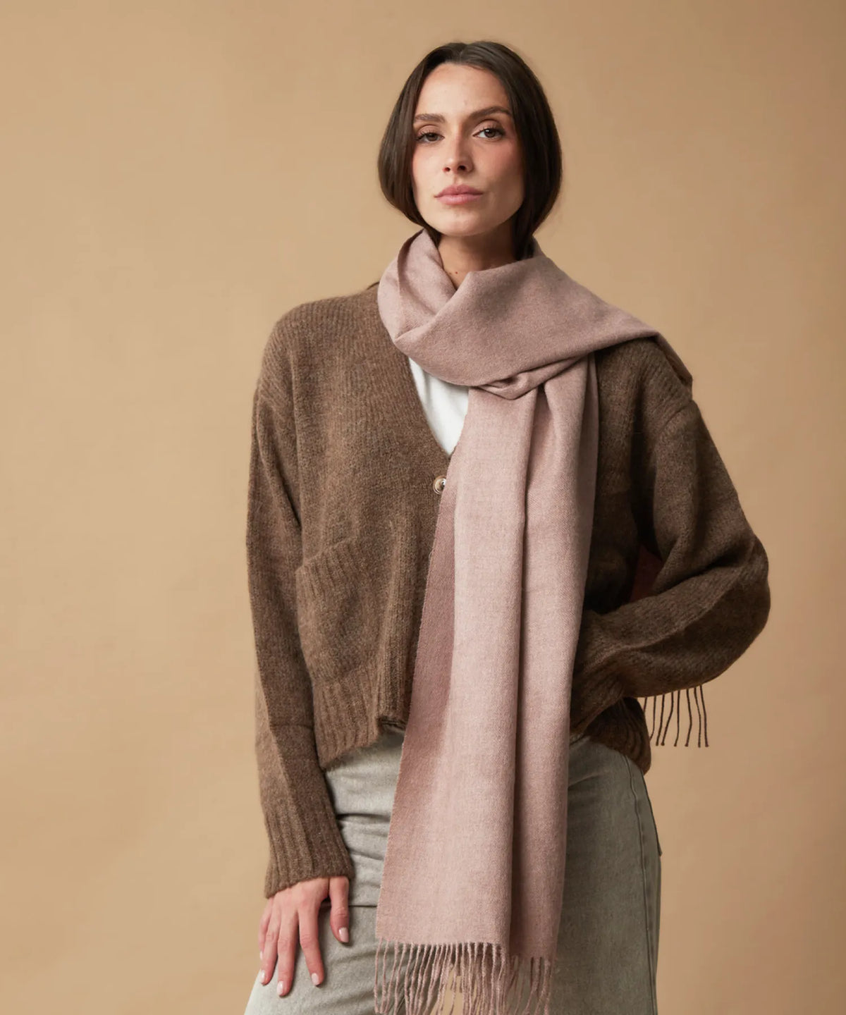 A woman wearing a brown knit cardigan, a light olive green skirt, and a dusty pink scarf, standing against a solid beige background.