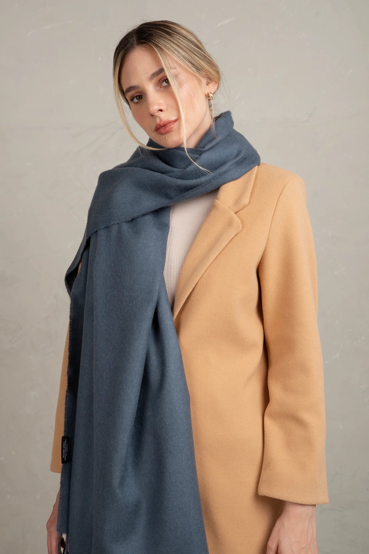 Woman wearing a beige coat and blue alpaca scarf against a plain background