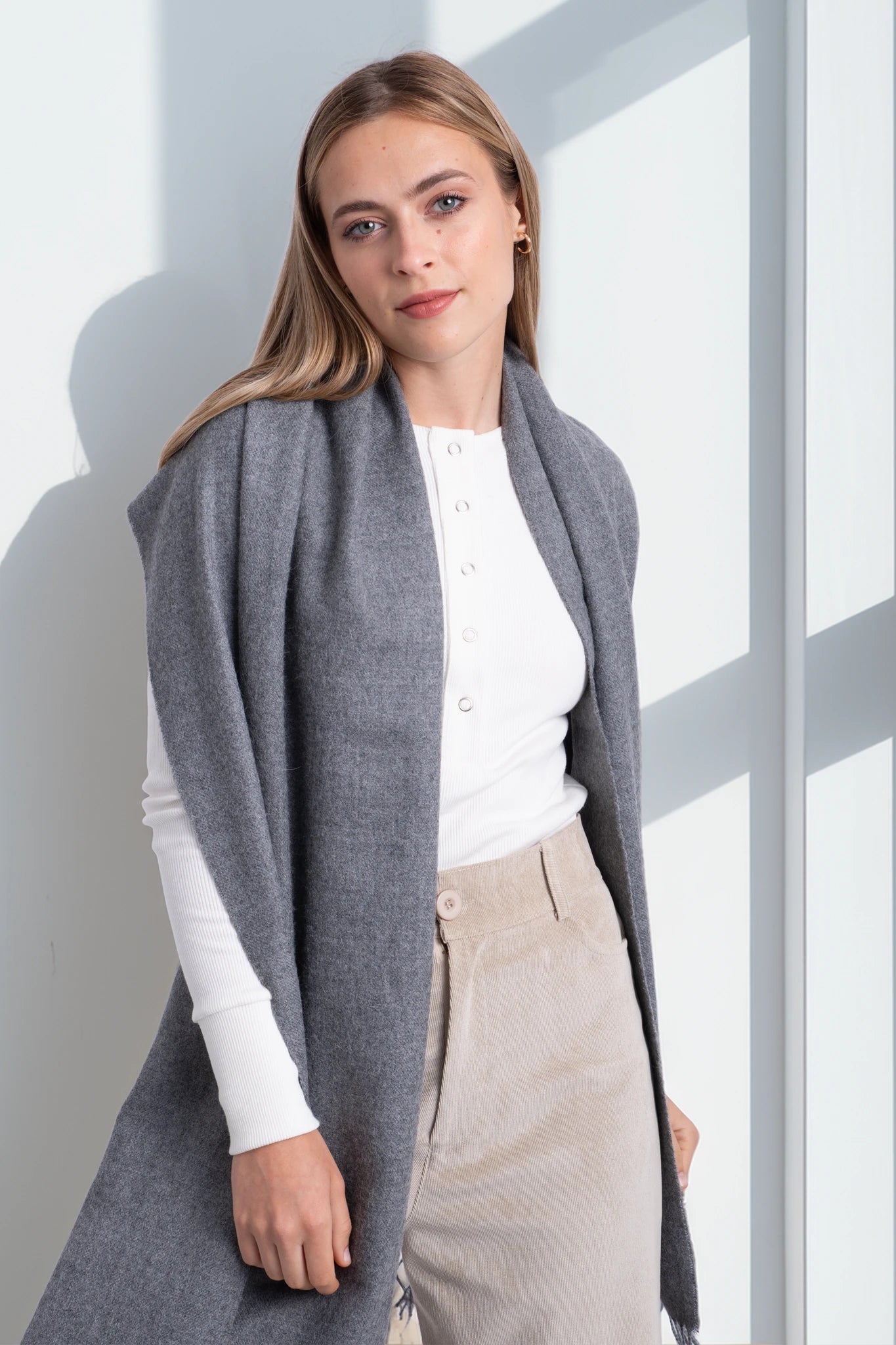 Woman wearing a gray cardigan over a white shirt and beige pants in a bright room.