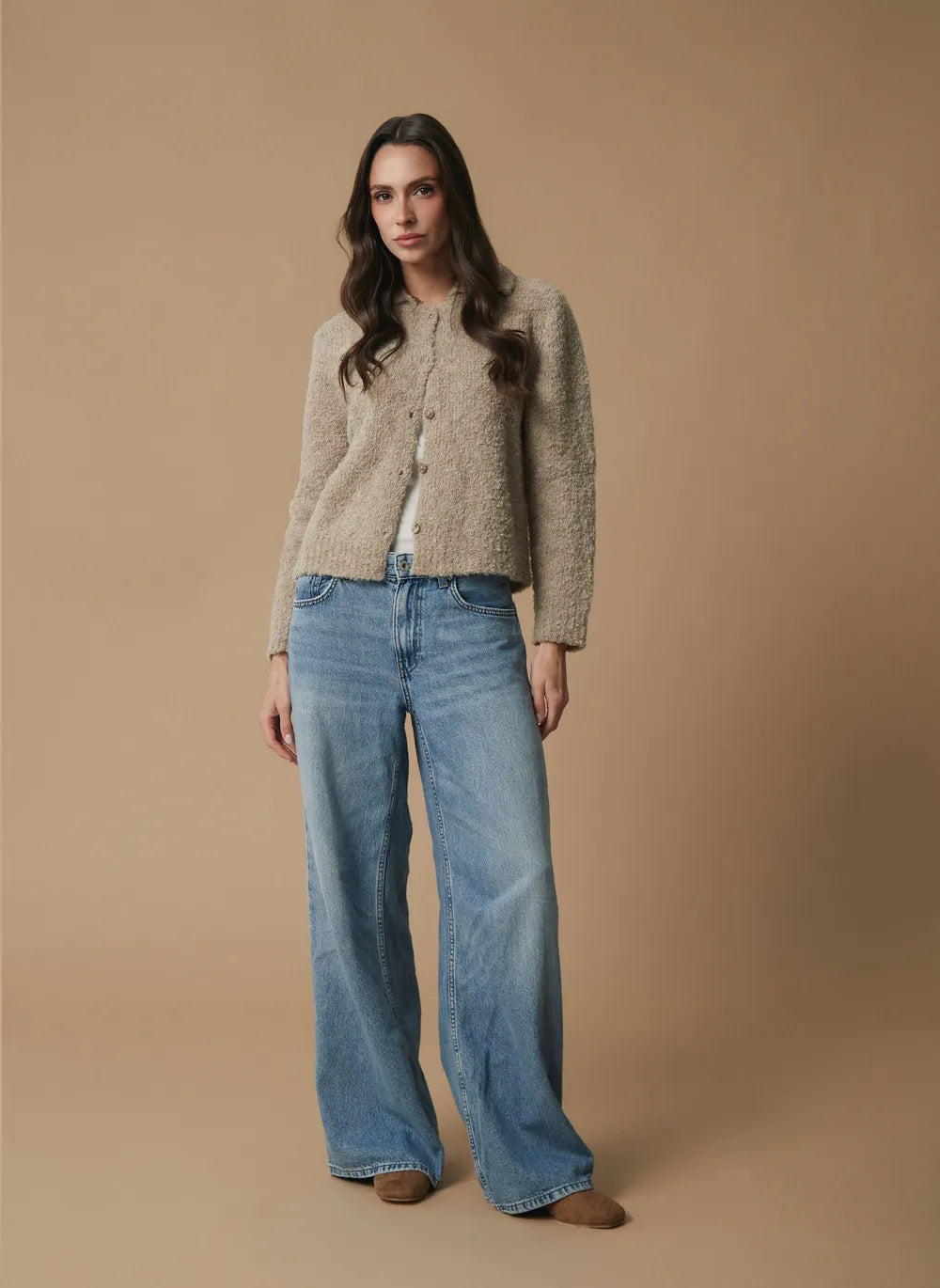 A woman stands against a plain warm beige studio background wearing a fuzzy cropped beige alpaca cardigan over a white top and wide-leg light blue jeans with brown shoes.
