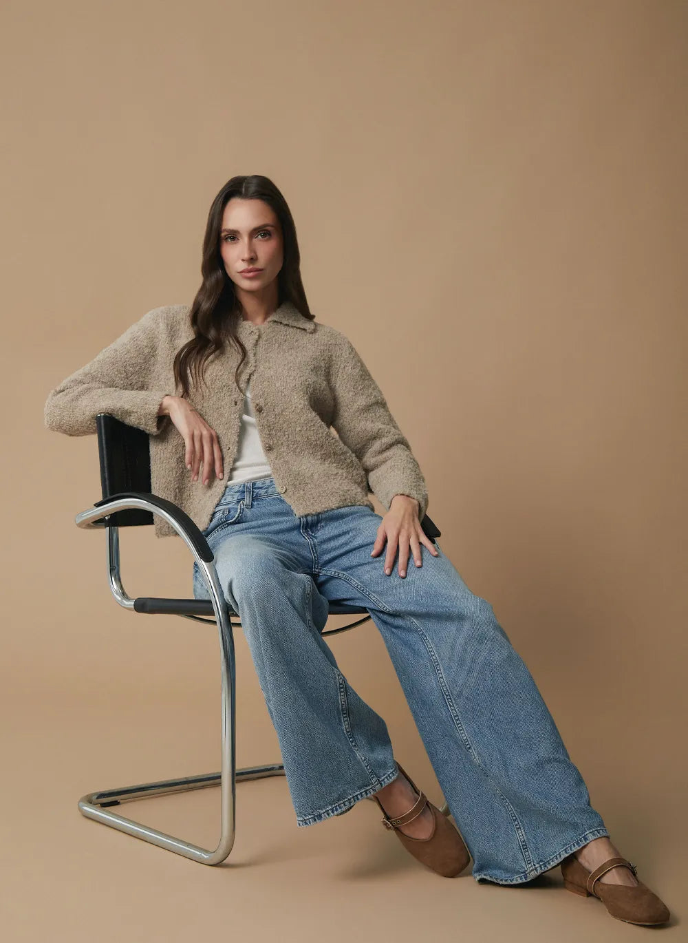 A woman sits on a modern metal chair against a solid beige studio background, wearing a fuzzy beige cardigan over a white top, wide-leg blue jeans, and brown flat shoes.