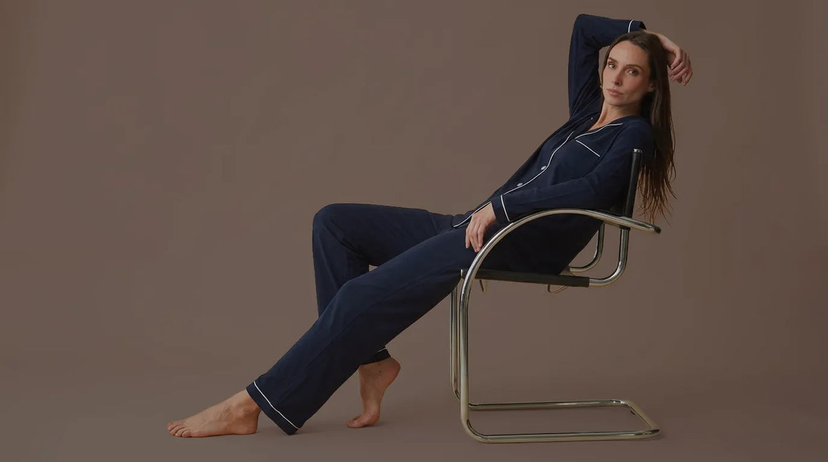 A woman wearing a navy blue pajama set with white piping sits relaxed on a modern metal chair against a solid taupe background.