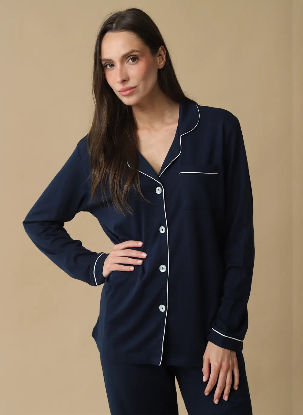 A woman modeling a navy blue pajama set with white piping details, consisting of a long-sleeve button-up top and matching pants, against a solid beige background.