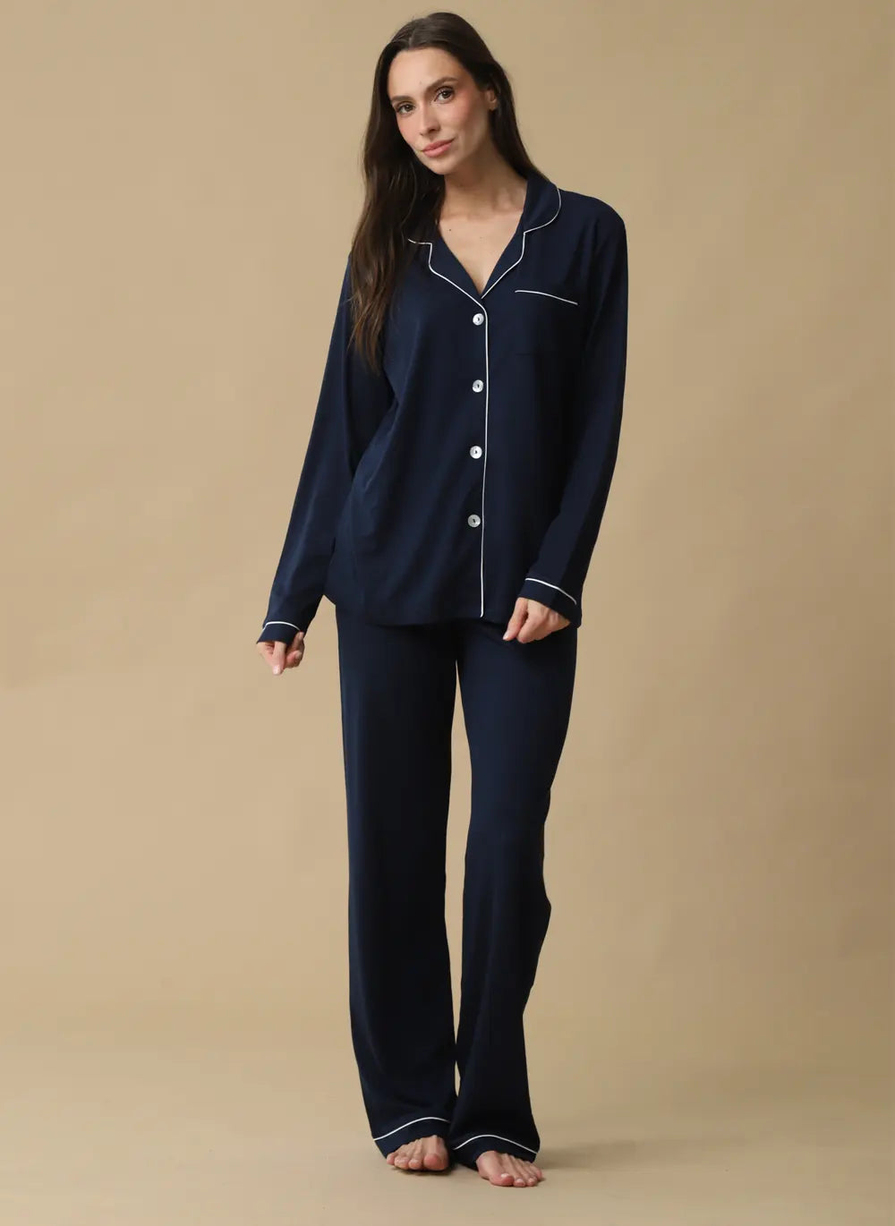 A woman modeling a two-piece navy blue pajama set with white piping details, consisting of a long-sleeve button-up top and matching pants, against a solid beige studio background.
