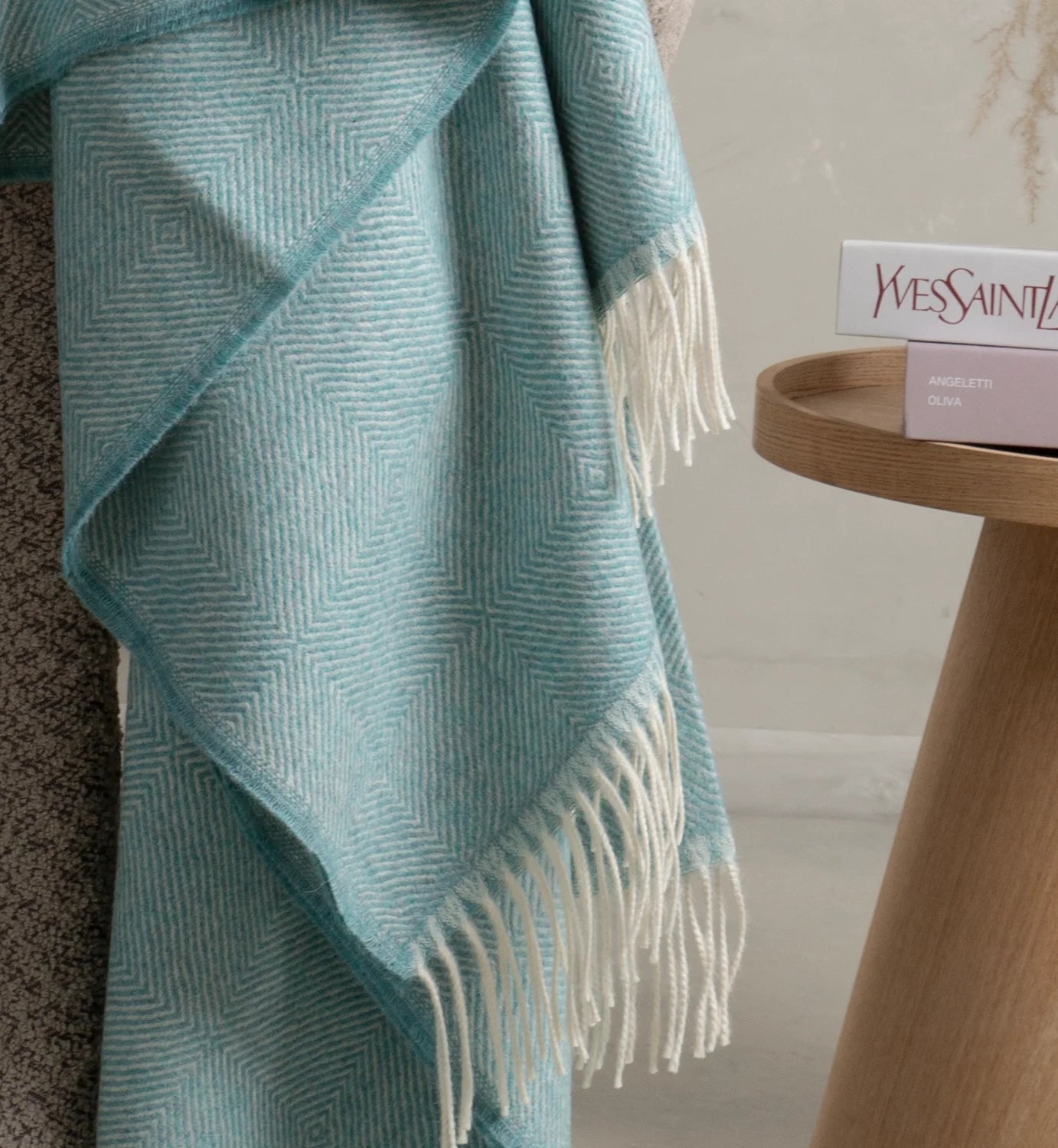 Teal patterned baby alpaca throw blanket draped over a chair next to a wooden side table with Yves Saint Laurent products.