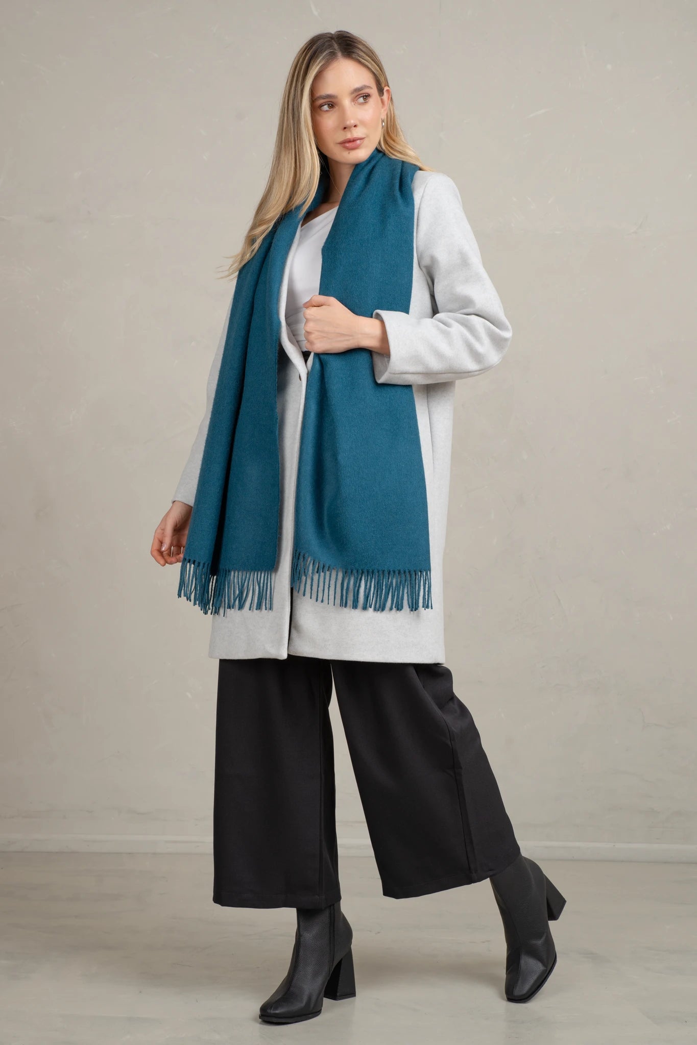 Woman wearing a blue alpaca scarf, white shirt, black pants, and black shoes against a plain background