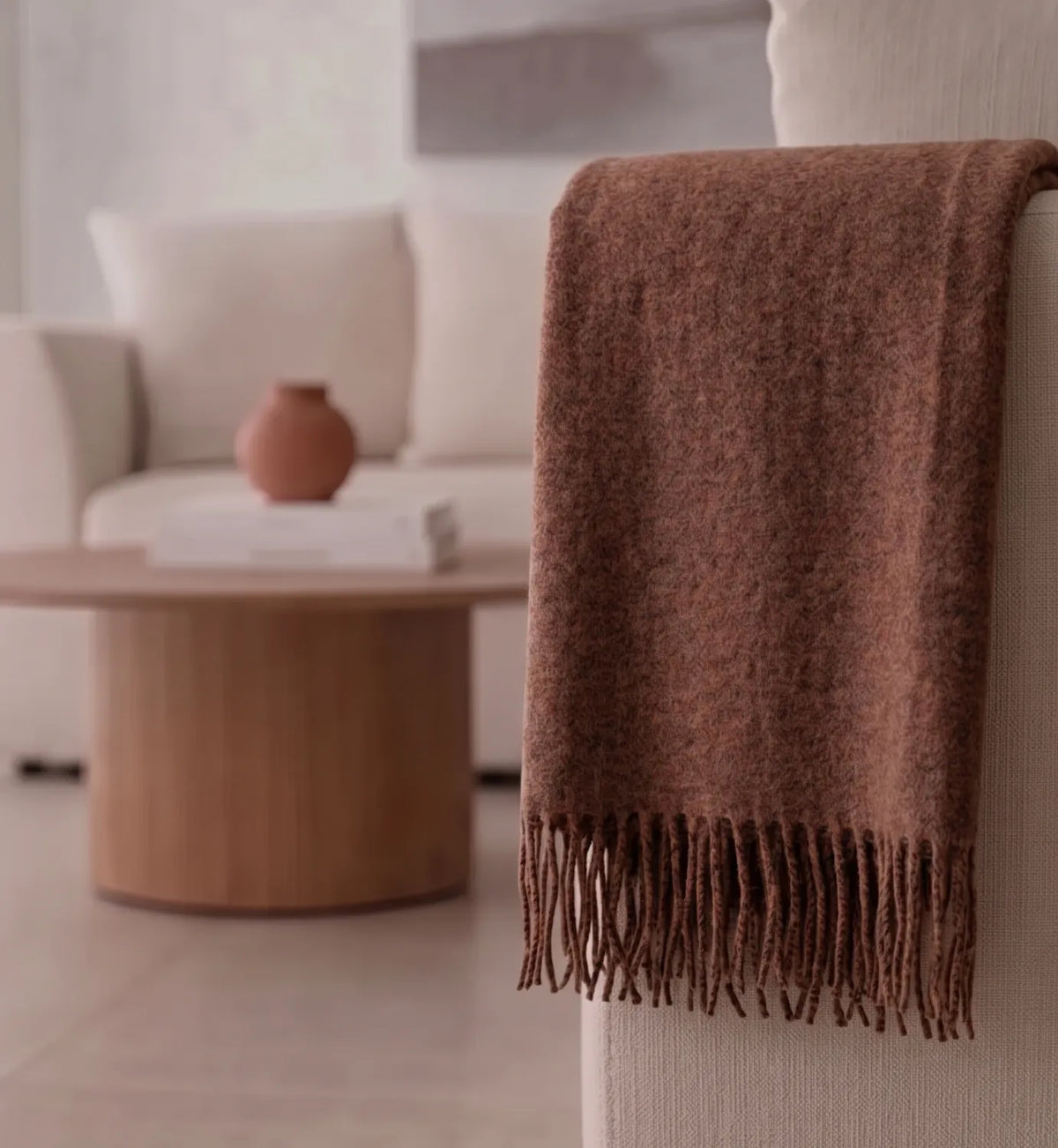 A warm brown textured throw blanket with fringe detailing draped over the arm of a cream-colored chair in a modern, softly lit living room.