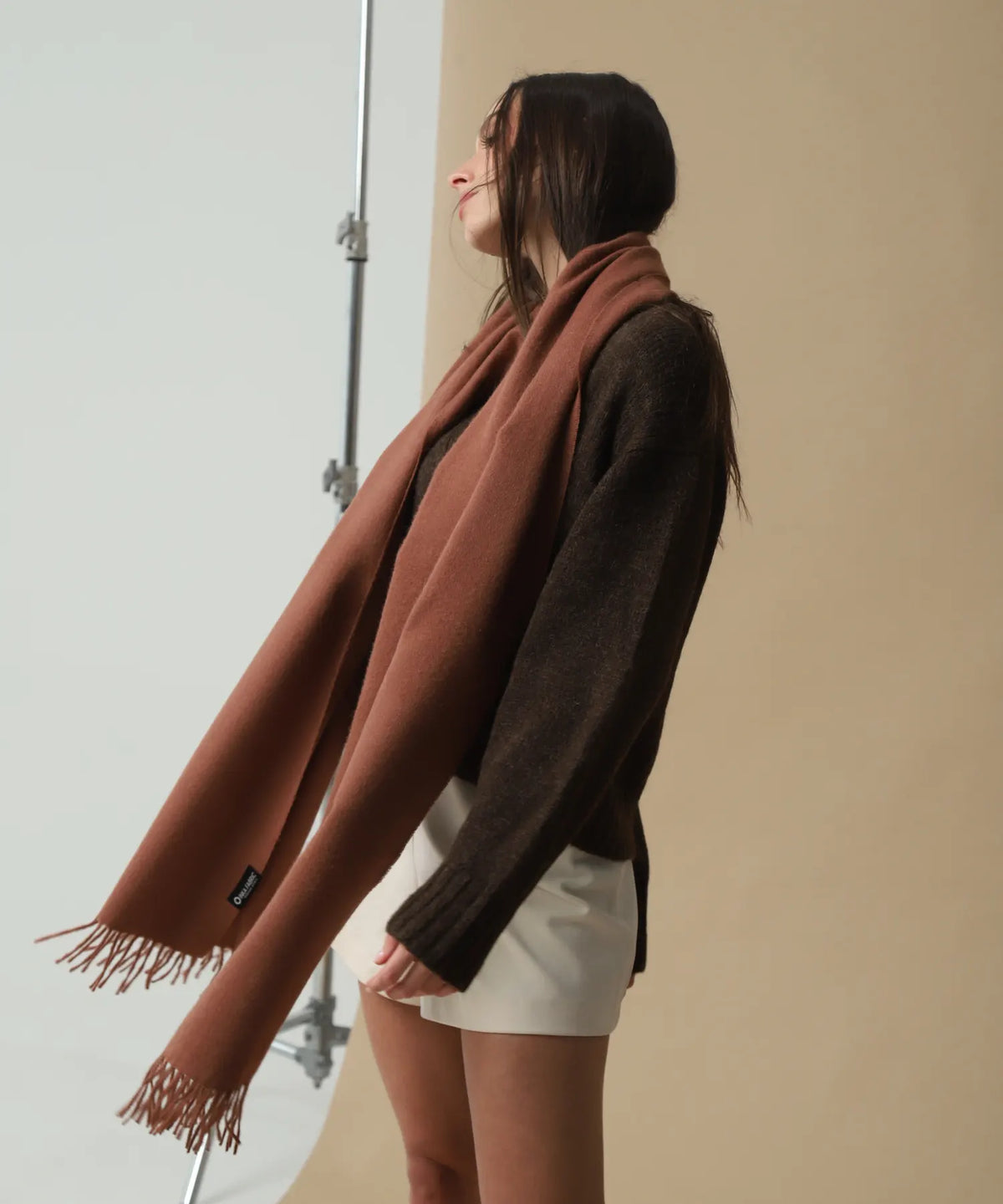 A woman wearing a long, warm brown scarf with fringe ends, styled over a dark brown sweater and white shorts, standing in a studio setting.