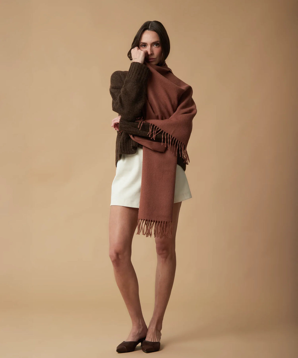 A woman models a rust-brown fringed shawl wrapped around her shoulders, styled over a dark brown knit sweater and white shorts against a solid beige studio background.