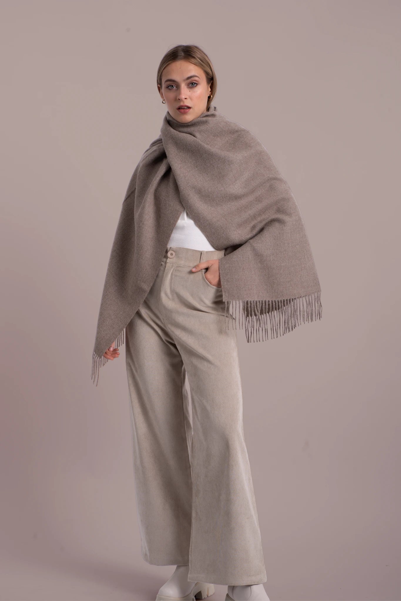 Woman wearing a beige scarf with fringe, white top, and beige pants against a plain background