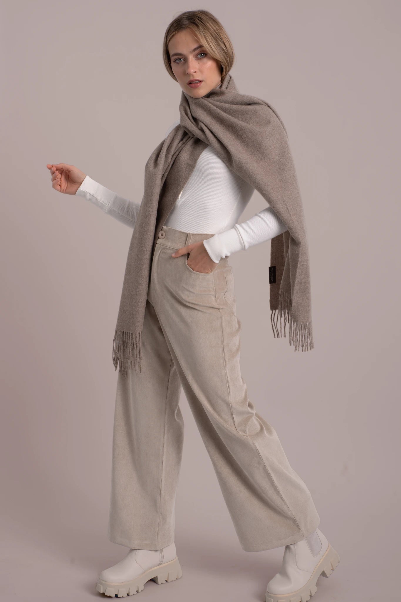 Woman wearing a beige scarf, white top, beige pants, and white shoes on a plain background