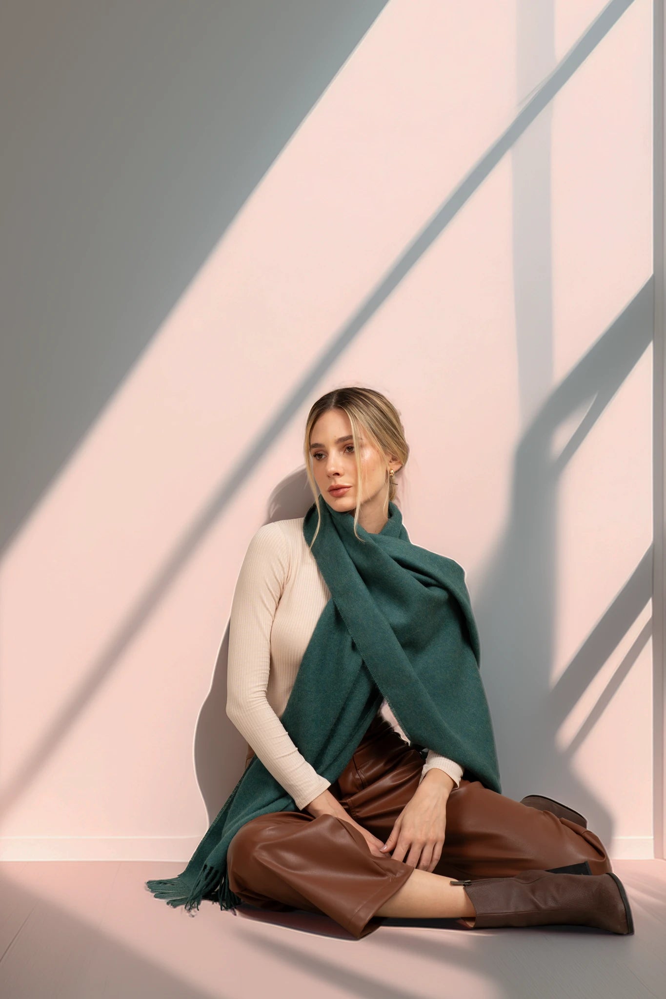 Woman sitting on a pink floor with a green scarf and brown pants, casting shadows on a light pink wall.