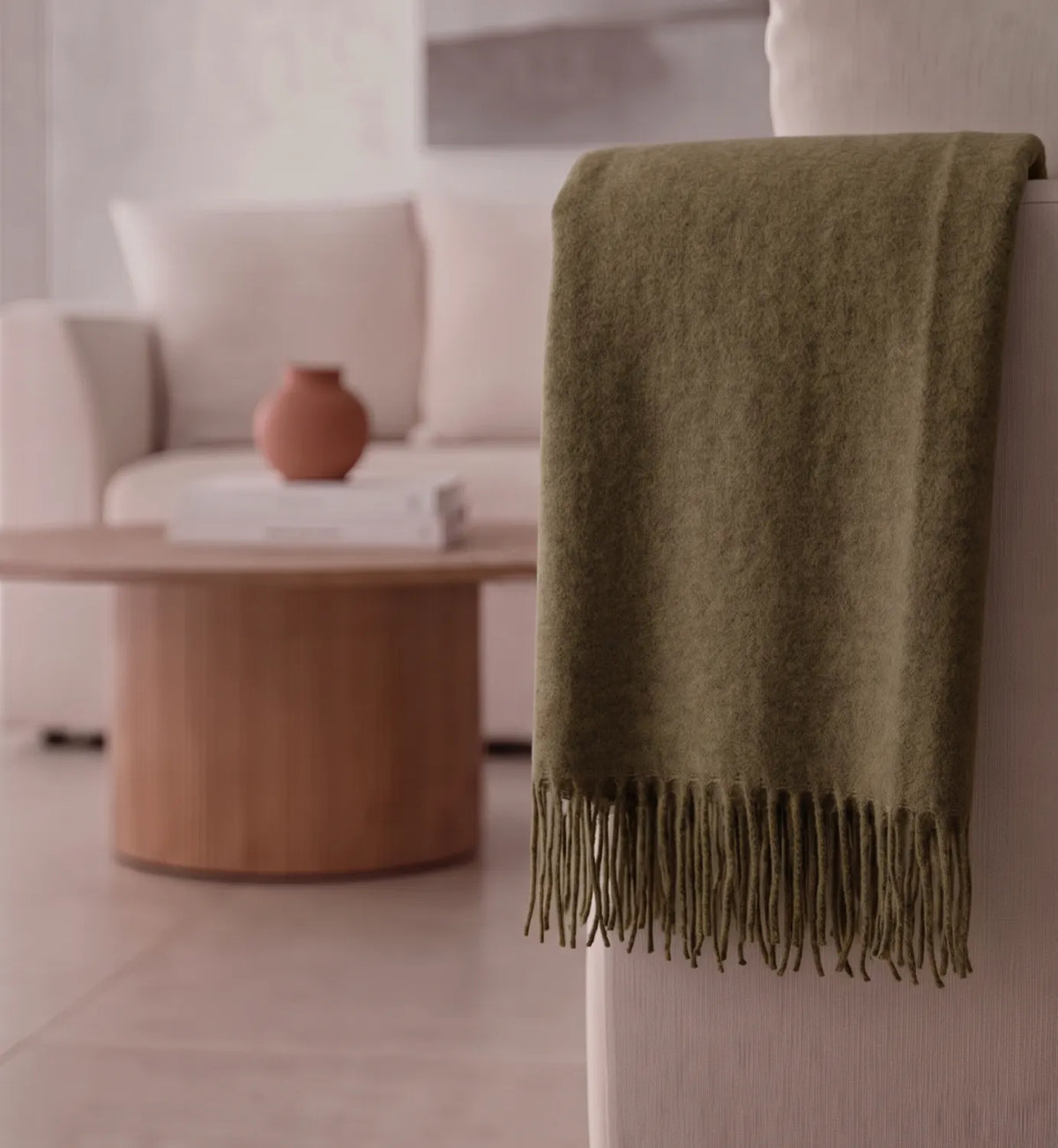 An olive green knit throw blanket with fringed edges draped over the back of a light-colored armchair in a softly lit living room.