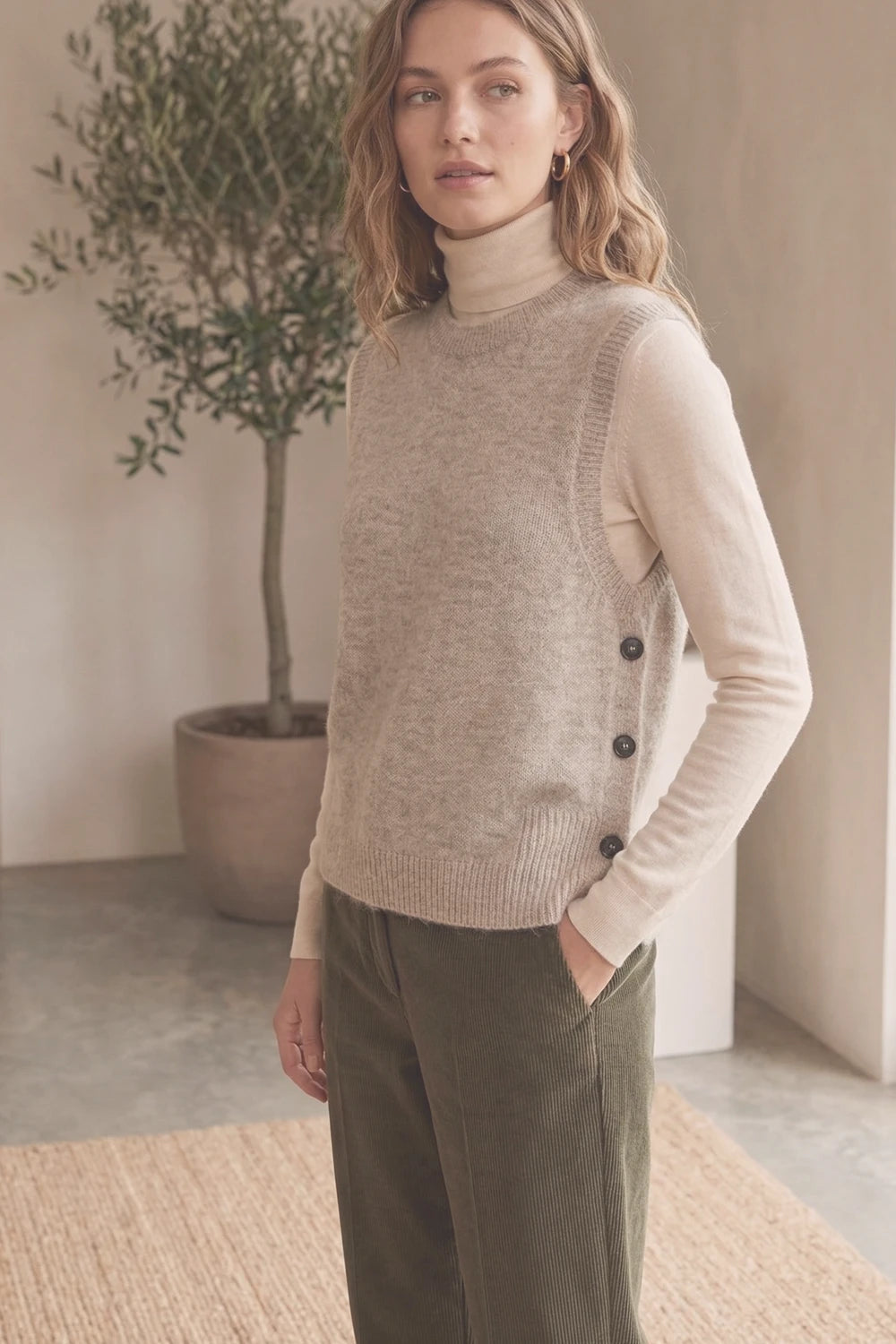 Woman wearing a beige sweater vest over a cream turtleneck with green pants, standing indoors.