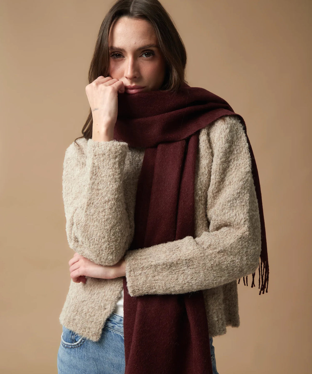 A woman modeling a chunky beige bouclé cardigan sweater and a deep burgundy scarf against a solid beige studio background.