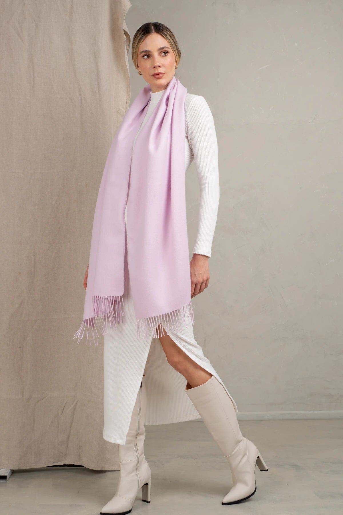 Woman wearing a alpaca light pink scarf and white outfit with knee-high boots against a neutral background