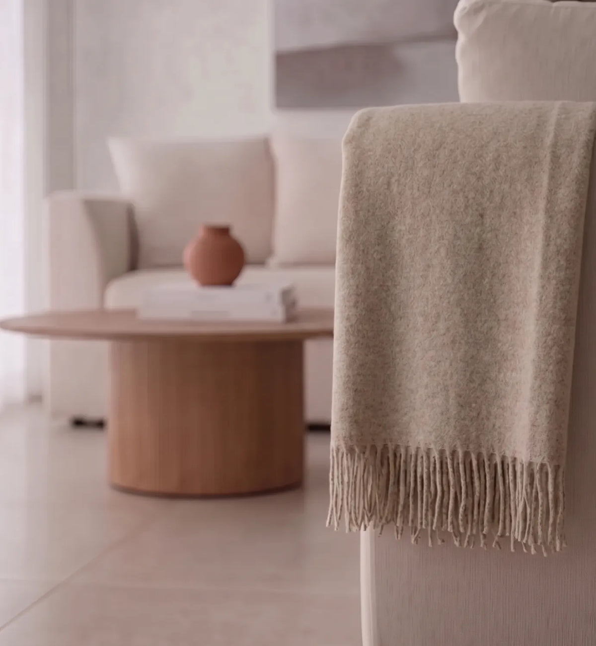 A soft beige throw blanket with fringe trim draped over the back of a light-colored armchair in a minimalist living room setting.