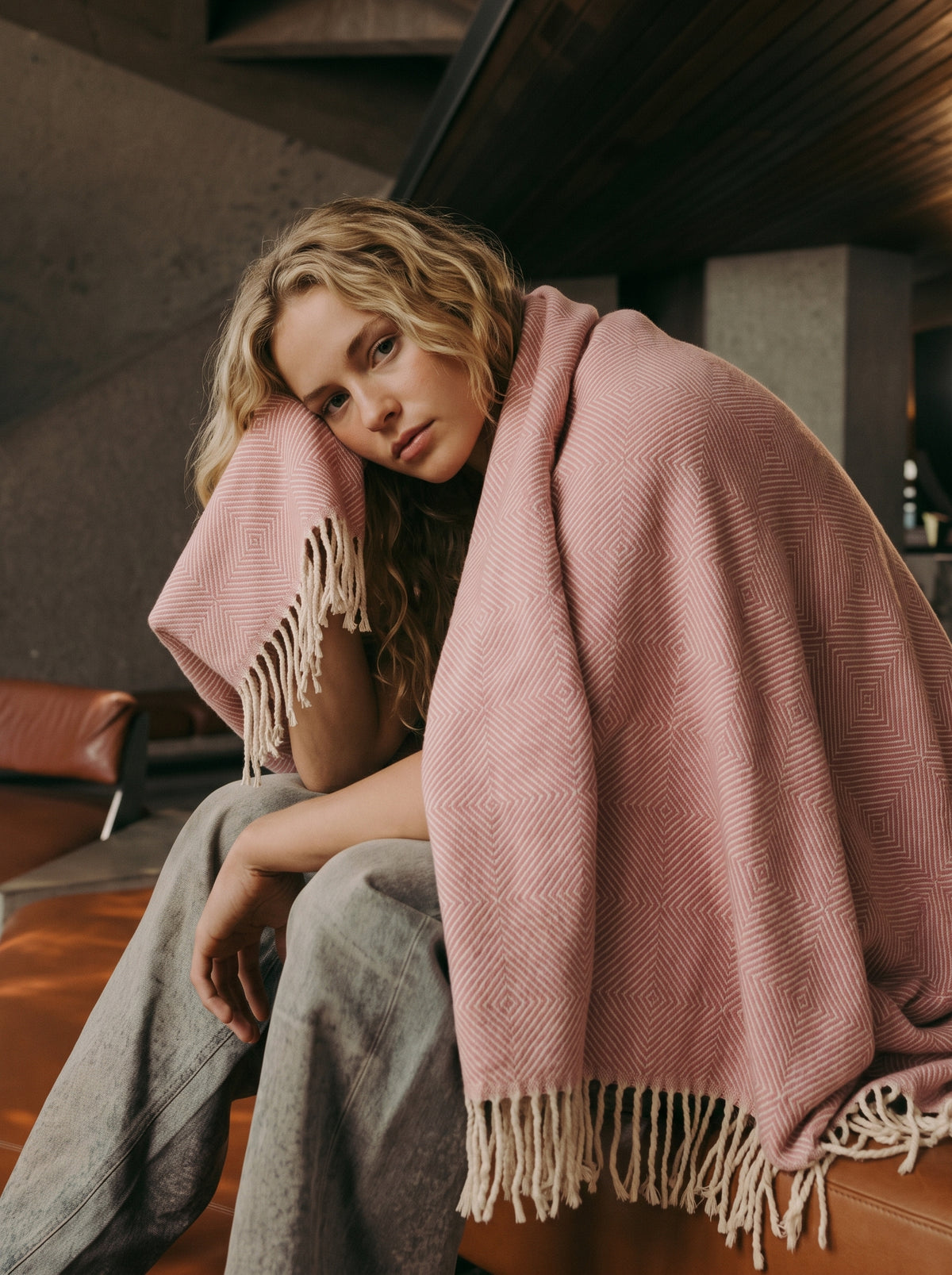 A woman sits wrapped in a dusty pink woven throw blanket with cream fringe, in a dimly lit indoor setting.