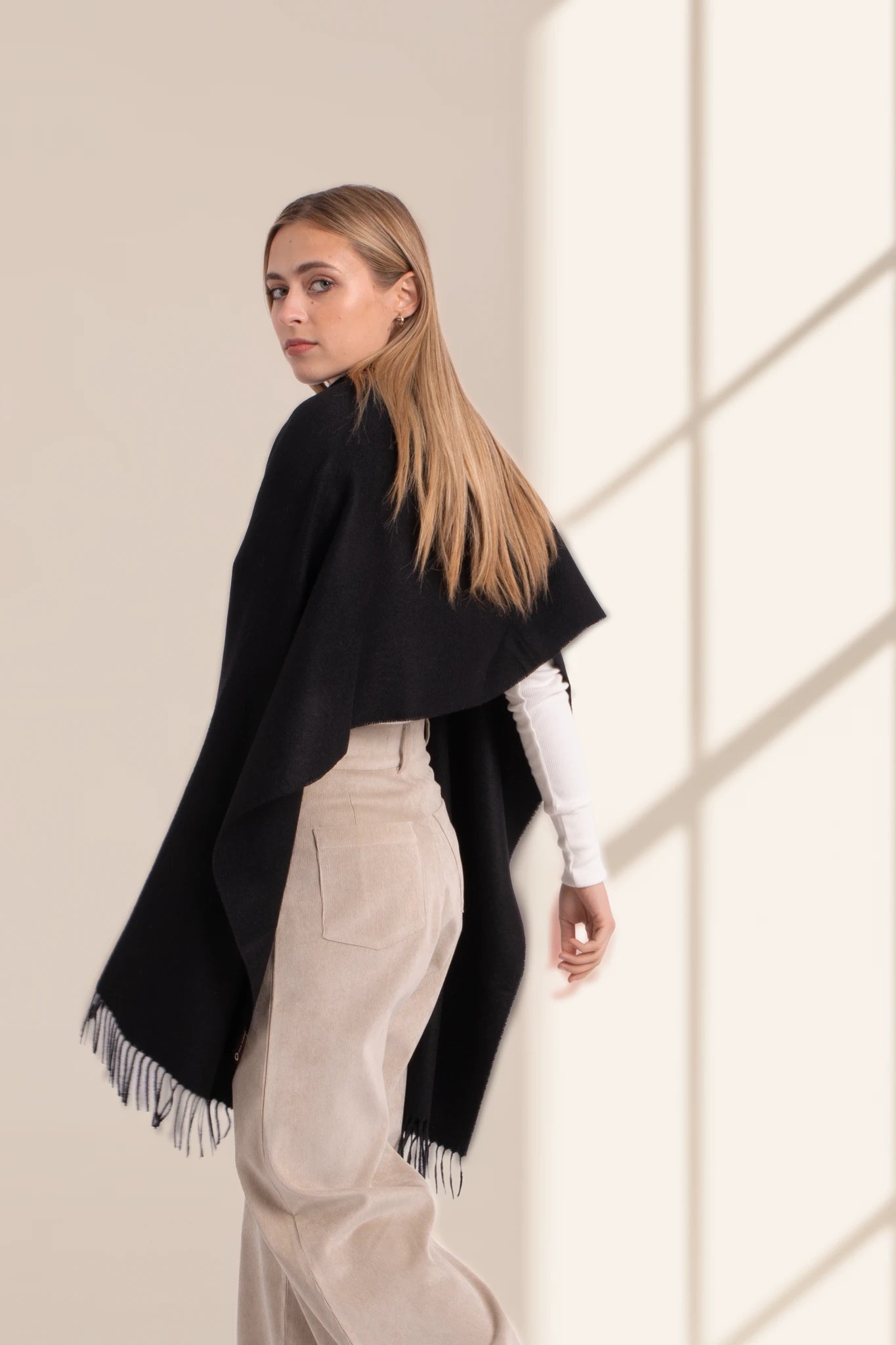 Woman wearing a black poncho with white pants against a neutral background