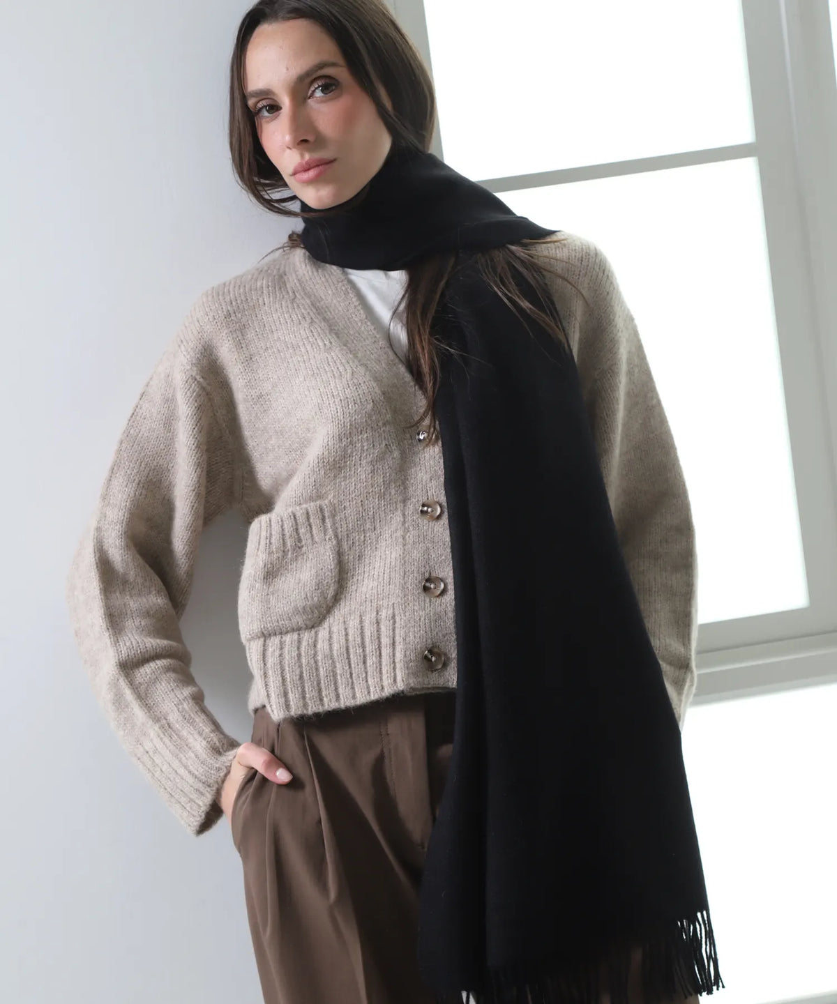 A woman wearing a beige knit cardigan, a black turtleneck, brown trousers, and a black scarf, standing in a bright room near a window.