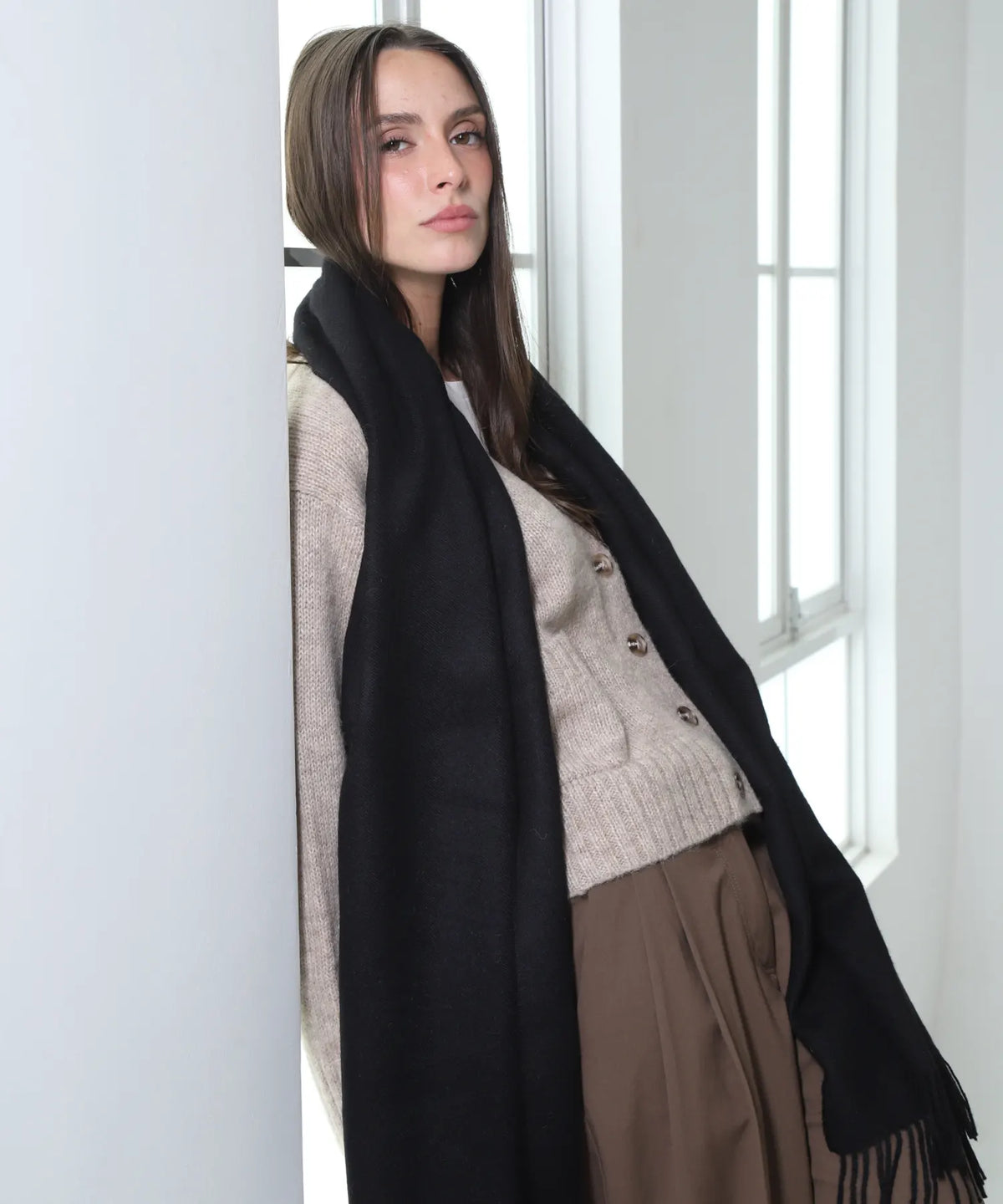 A woman wearing a thick black scarf draped over a beige cardigan and brown skirt, standing in a brightly lit room next to a window.