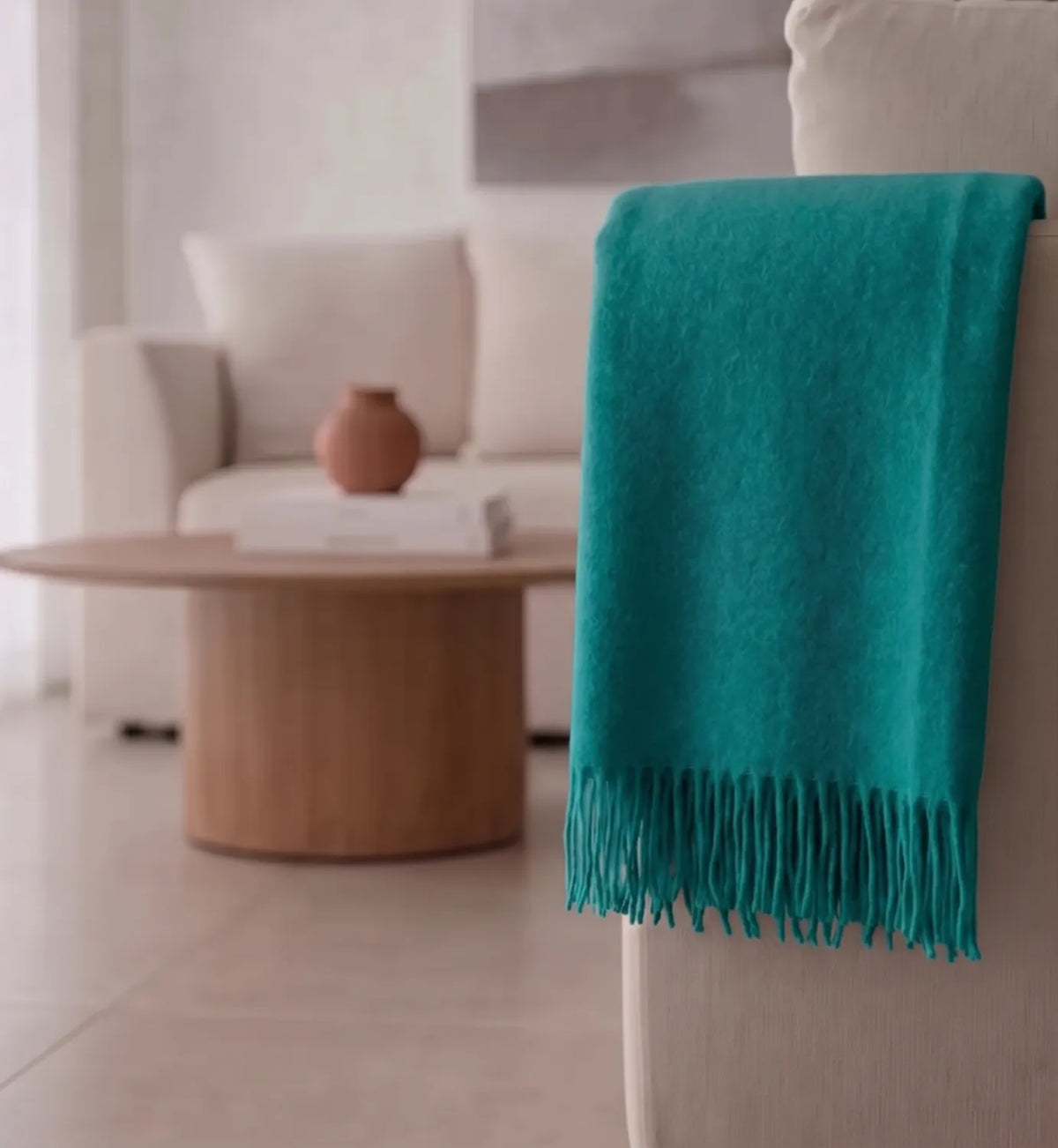 A teal throw blanket with fringe trim draped over the arm of a light-colored sofa in a softly lit living room.