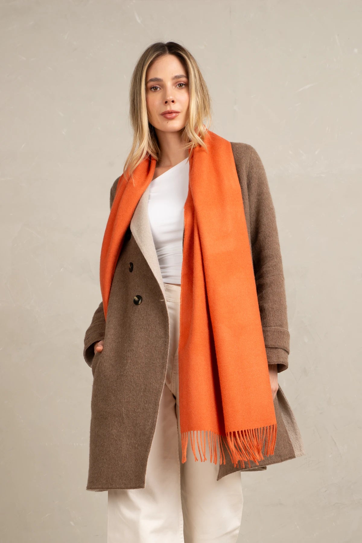Woman wearing a brown coat and alpaca scarf orange scarf against a beige background