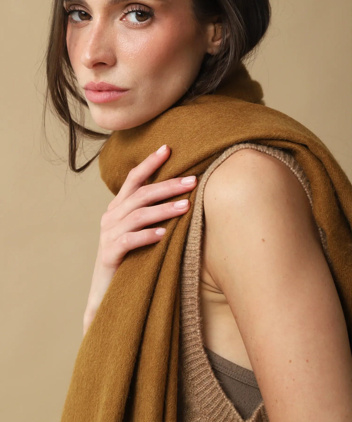 A close-up lifestyle shot of a model wearing a warm camel brown knit sweater vest and a matching camel brown knit scarf, set against a solid beige background.