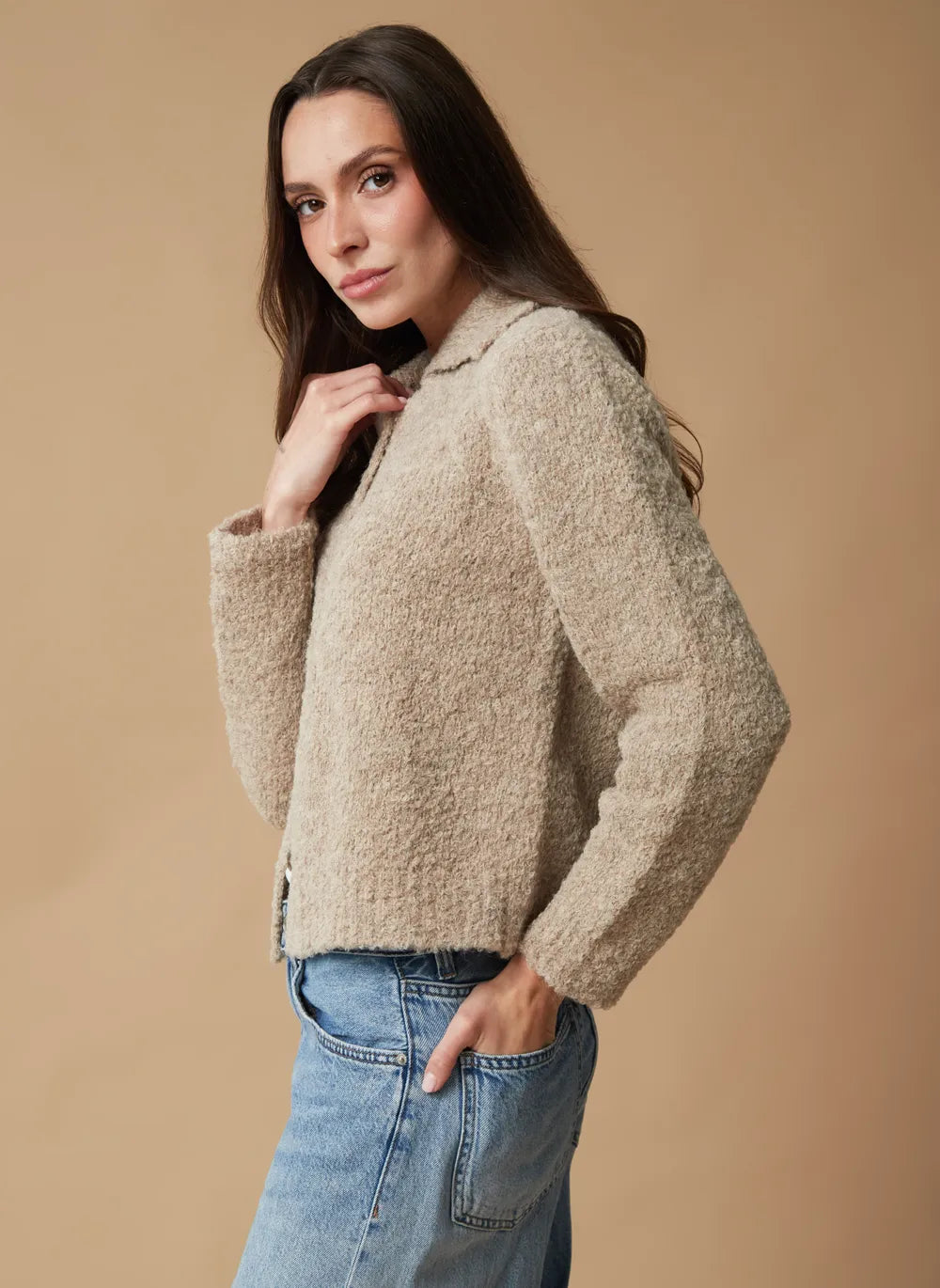 A woman wearing a textured beige cropped cardigan sweater and blue jeans, posing against a solid warm beige studio background.