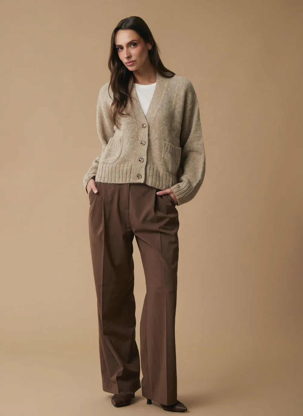 A woman modeling a beige knit cardigan over a white top and brown wide-leg trousers, standing against a solid beige studio background.