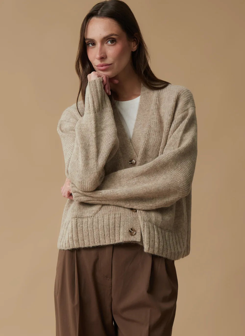 A woman wearing a beige ribbed knit cardigan over a white top and brown trousers, posed against a solid beige studio background.